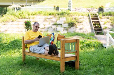 Parkta bankta oturan ve gümüş dizüstü bilgisayar kullanırken cep telefonuyla konuşan 50 'li yaşlarda Avrupalı bir adamın görüntüsü