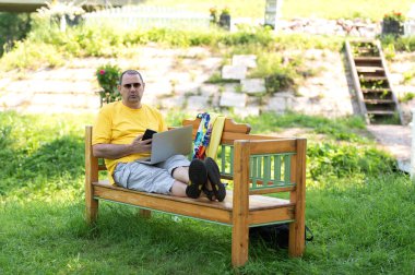 Parkta bankta oturan ve gümüş dizüstü bilgisayar kullanırken cep telefonuyla konuşan 50 'li yaşlarda Avrupalı bir adamın görüntüsü