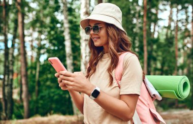 Şapkalı ve sırt çantalı bir kadın yürüyüşçü ormanda bir telefonla bekliyor. Kamp, seyahat, yürüyüş..