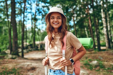 Şapkalı ve sırt çantalı bir kadın yürüyüşçü ormanın arka planında duruyor. Kamp, seyahat, yürüyüş..