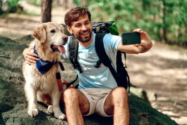 Sırt çantası ve labrador köpeği olan bir erkek yürüyüşçü ormanda bir taşın üzerinde otururken telefonda selfie çekiyor. Kamp, seyahat, yürüyüş..