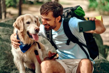 Sırt çantası ve labrador köpeği olan bir erkek yürüyüşçü ormanda bir taşın üzerinde otururken telefonda selfie çekiyor. Kamp, seyahat, yürüyüş..