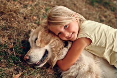 Sevimli küçük sarışın bir kız çocuğu çimenlerde uzanıyor arkadaşına sarılıyor labrador bir köpeğe.