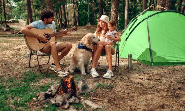 Çocuklu genç bir aile ve labrador köpeği çadırın yanındaki kamp ateşinin yanında oturup çam ormanında gitar çalıyorlar. Kamp, dinlenme, yürüyüş..