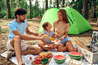 Mutlu bir aile ve piknik yapan bir çocuk çadırın yakınındaki bir battaniyede oturuyor ve hafta sonu ormanda kazıkta kızartılmış yiyecekler yiyor. Kamp, dinlenme, yürüyüş..