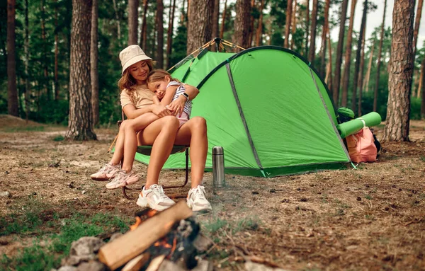 A young mother sits with her daughter near a campfire near a tent in a pine forest. Camping, recreation, hiking.