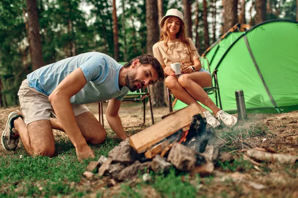 A young couple are sitting by a campfire near the tent, drinking coffee in a pine forest. Camping, recreation, hiking.