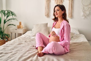 Genç hamile kadın yatak odasında yatakta otururken ellerini nazikçe karnının üstünde tutuyor. Annelik kavramı.