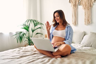 Genç hamile bir kadın yatak odasında oturmuş video konuşmasında el sallıyor. Annelik kavramı.