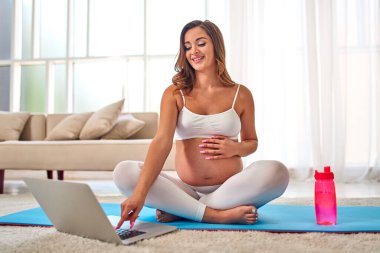Spor kıyafetli genç hamile bir kadın oturma odasındaki lastik paspasın üzerinde egzersiz yapıyor ve video derslerinde dizüstü bilgisayarı var. Hamilelik boyunca spor ve sağlıklı bir yaşam tarzı..
