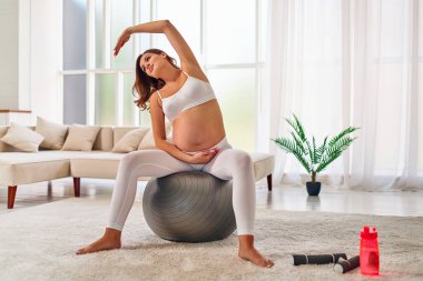 Spor giyimli genç hamile bir kadın spor salonunda spor yaparken spor topunun üzerinde oturuyor. Hamilelik boyunca spor ve sağlıklı bir yaşam tarzı..