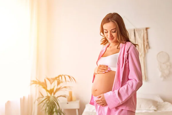 一位年轻的孕妇站在靠近窗户的卧室里 双手轻轻地放在肚子上 怀孕的概念 — 图库照片