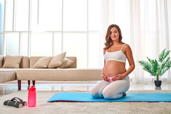 穿着运动服的年轻孕妇在客厅的橡胶垫子上做运动 附近有哑铃和一瓶水 怀孕期间的运动和健康生活方式 — 图库照片