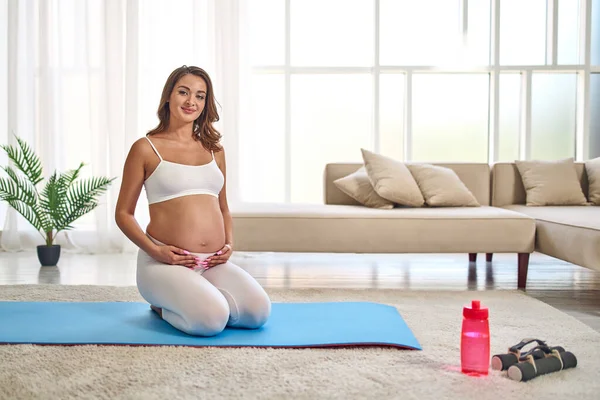 穿着运动服的年轻孕妇在客厅的橡胶垫子上做运动 附近有哑铃和一瓶水 怀孕期间的运动和健康生活方式 — 图库照片