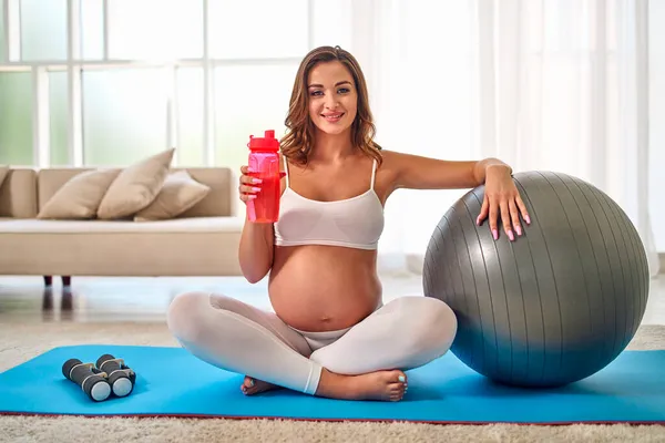 身穿运动服的年轻孕妇在客厅里用橡皮垫上的球做运动 怀孕期间的运动和健康生活方式 — 图库照片