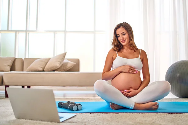 身穿运动服的年轻孕妇穿着运动服 带着笔记本电脑在客厅的橡胶垫子上做运动 怀孕期间的运动和健康生活方式 — 图库照片