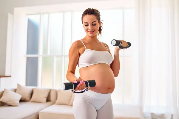 穿着运动服的年轻孕妇在客厅里用哑铃做运动 怀孕期间的运动和健康生活方式 — 图库照片