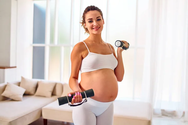 穿着运动服的年轻孕妇在客厅里用哑铃做运动 怀孕期间的运动和健康生活方式 — 图库照片