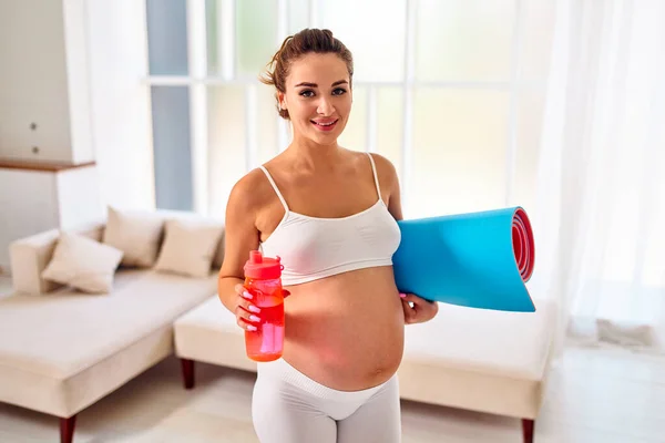 身穿运动服的年轻孕妇拿着一个橡胶垫子 从客厅的瓶子里喝水 怀孕期间的运动和健康生活方式 — 图库照片