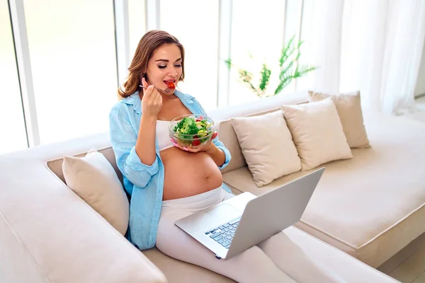 年轻的孕妇坐在沙发上 带着笔记本电脑和一盘新鲜的沙拉 健康饮食 工作从家里 自由职业 网上购物 产妇概念 — 图库照片