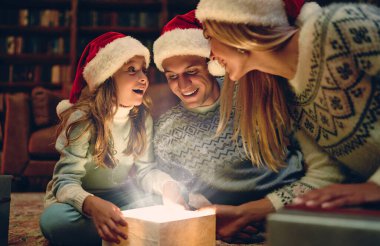 Mutlu Noeller ve mutlu yıllar, mutlu aile Noel Baba şapkalarıyla yeni yılı bekliyor. Aileler sevimli kızlarına hediye paketi sunuyorlar. İçeriden sihirli ışık.
