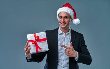 Attractive successful businessman in a jacket and shirt in a cap of Santa Claus with a gift box in the studio on a gray background.