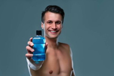 Handsome young man isolated. Portrait of shirtless muscular man is standing on grey background with bottle of mouthwash. Men care concept.