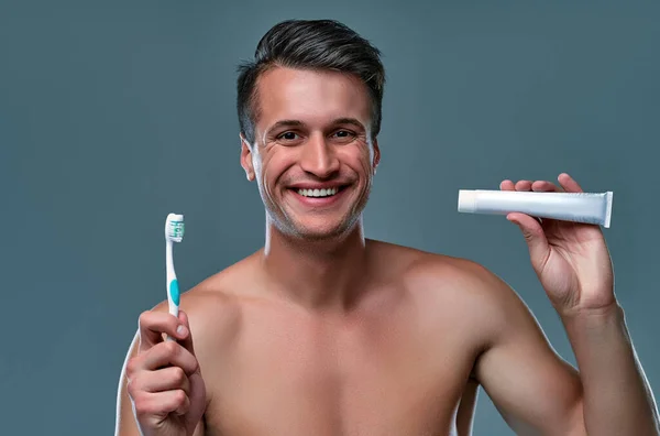 ハンサムな若い男隔離 上半身裸の筋肉男性の肖像画は手に歯ブラシと歯磨き粉と灰色の背景に立っています 男性ケア概念 — ストック写真