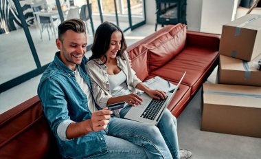 Genç çift yeni eve taşınırken bilgisayar kablosuz internet ve kredi kartı kullanarak yerde birlikte oturuyorlar. Taşınmak, ev almak, apartman konsepti..