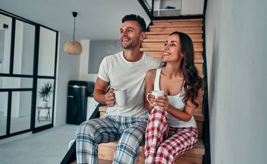 Pijama giymiş genç bir çift evlerinin merdivenlerinde oturmuş sabah kahvelerini yudumluyorlar..