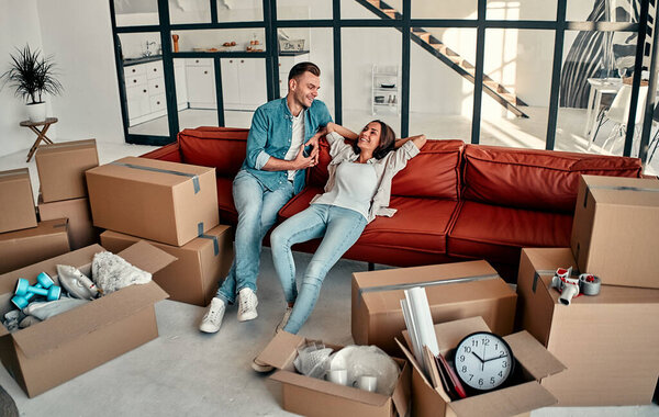Young married couple sitting on the sofa in the living room at home. Happy husband and wife are having fun, are looking forward to a new home. Moving and relocate new home concept.