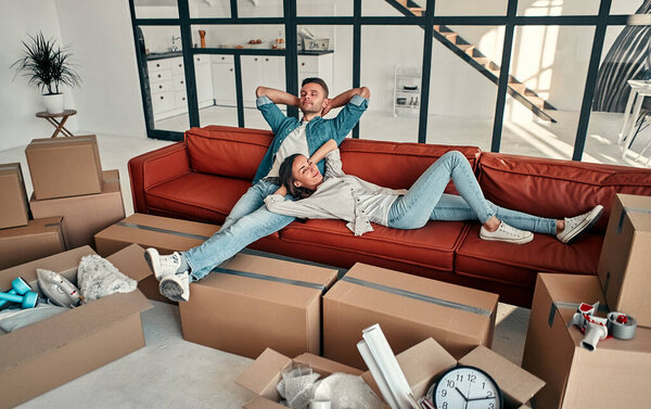 Young married couple sitting on the sofa in the living room at home. Happy husband and wife are having fun, are looking forward to a new home. Moving, buying a house, apartment concept.