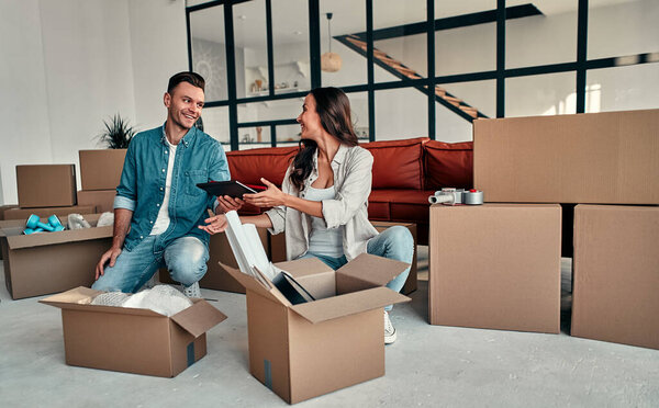 A young married couple unpacking things from boxes in the living room in the house. Happy husband and wife are having fun, are looking forward to a new home. Moving, buying a house, apartment concept.