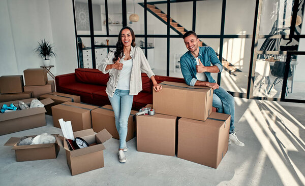 A young married couple in the living room in the house stand near unpacked boxes. Happy husband and wife show thumbs up, are happy about new home. Moving, buying a house, apartment concept.
