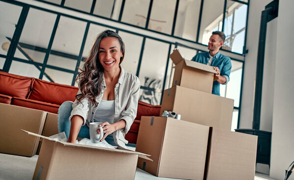 A young married couple in the living room in the house unpack boxes with things. Happy husband and wife are having fun, are looking forward to a new home. Moving, buying a house, apartment concept.