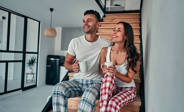 パジャマを着た若いカップルが家の階段に座って朝のコーヒーを飲んでいます — ストック写真