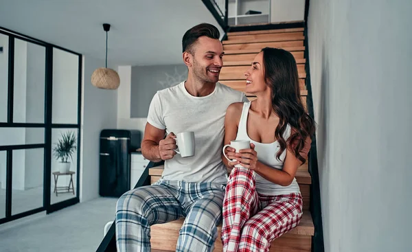 パジャマを着た若いカップルが家の階段に座って朝のコーヒーを飲んでいます — ストック写真