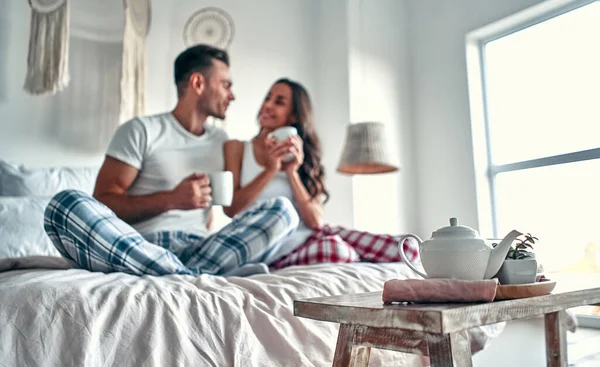 ベッドの上に座って家で朝コーヒーを飲んでいる間 幸せな若いカップルが話して笑っています — ストック写真