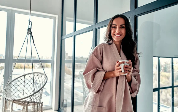 Young woman chilling at home against the background of a window and a hanging chair with a cup of coffee in hand. Girl relaxing in living room wearing home bathrobe.