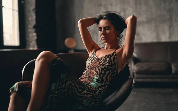一个漂亮的黑发美女的画像 穿着一件坐在扶手椅上摆姿势的连衣裙 用手把她长长的卷发直 — 图库照片