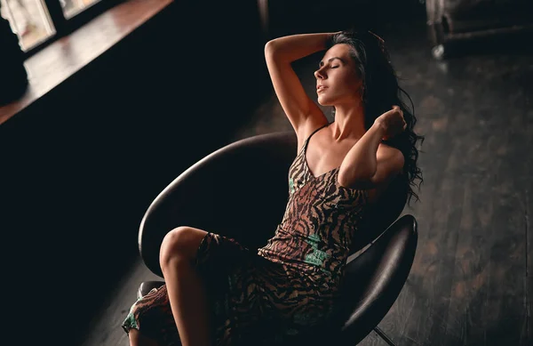 一个穿着连衣裙 坐在扶手椅上摆姿势 用手把她长长的卷发直的美丽的黑发的画像 — 图库照片
