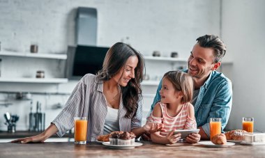Mutfakta mutlu aile kavramı. Şirin küçük kız ve güzel ailesi akıllı bir telefon kullanıyor ve lezzetli bir kahvaltı hazırlarken gülümsüyorlar..
