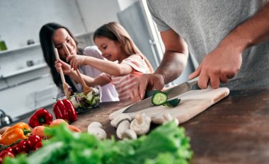 Anne, baba ve kızı mutfakta yemek pişiriyor. Mutlu aile kavramı. Yakışıklı adam, çekici genç kadın ve tatlı küçük kızları birlikte salata yapıyorlar. Sağlıklı yaşam tarzı.