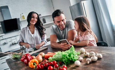 Anne, baba ve kızı mutfakta yemek pişiriyor. Mutlu aile kavramı. Yakışıklı adam, çekici genç kadın ve tatlı kızları salata yapıyorlar ve akıllı telefon kullanıyorlar..
