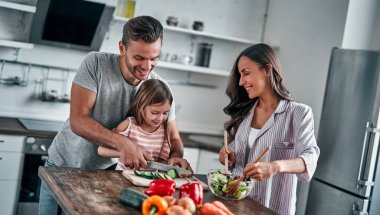 Anne, baba ve kızı mutfakta yemek pişiriyor. Mutlu aile kavramı. Yakışıklı adam, çekici genç kadın ve tatlı küçük kızları birlikte salata yapıyorlar. Sağlıklı yaşam tarzı.
