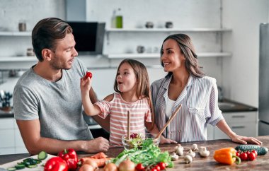 Anne, baba ve kızı mutfakta yemek pişiriyor. Mutlu aile kavramı. Yakışıklı adam, çekici genç kadın ve tatlı küçük kızları birlikte salata yapıyorlar. Sağlıklı yaşam tarzı.