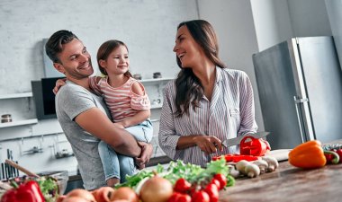 Anne, baba ve kızı mutfakta yemek pişiriyor. Mutlu aile kavramı. Yakışıklı adam, çekici genç kadın ve tatlı küçük kızları birlikte salata yapıyorlar. Sağlıklı yaşam tarzı.