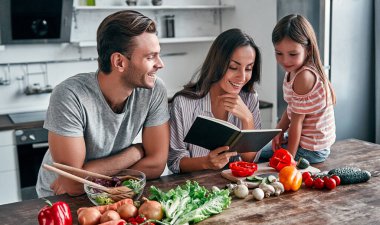 Anne, baba ve kızı yemek tarifi kitabıyla mutfakta yemek pişiriyorlar. Mutlu aile kavramı. Yakışıklı adam, çekici genç kadın ve tatlı küçük kızları birlikte salata yapıyorlar. Sağlıklı yaşam tarzı.