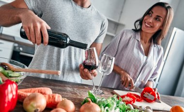 Romantik çift mutfakta yemek pişiriyor. Yakışıklı adam bardaklara şarap dolduruyor ve çekici genç bayan salata yaparken birlikte eğleniyor. Sağlıklı yaşam tarzı kavramı.