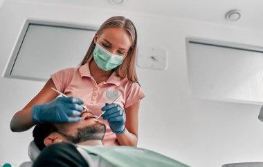 Diş kliniğinde aletleri olan bir hastayı maskeli kadın diş hekimi muayene ediyor. Doktor diş hekiminin sandalyesinde diş tedavisi yapıyor..